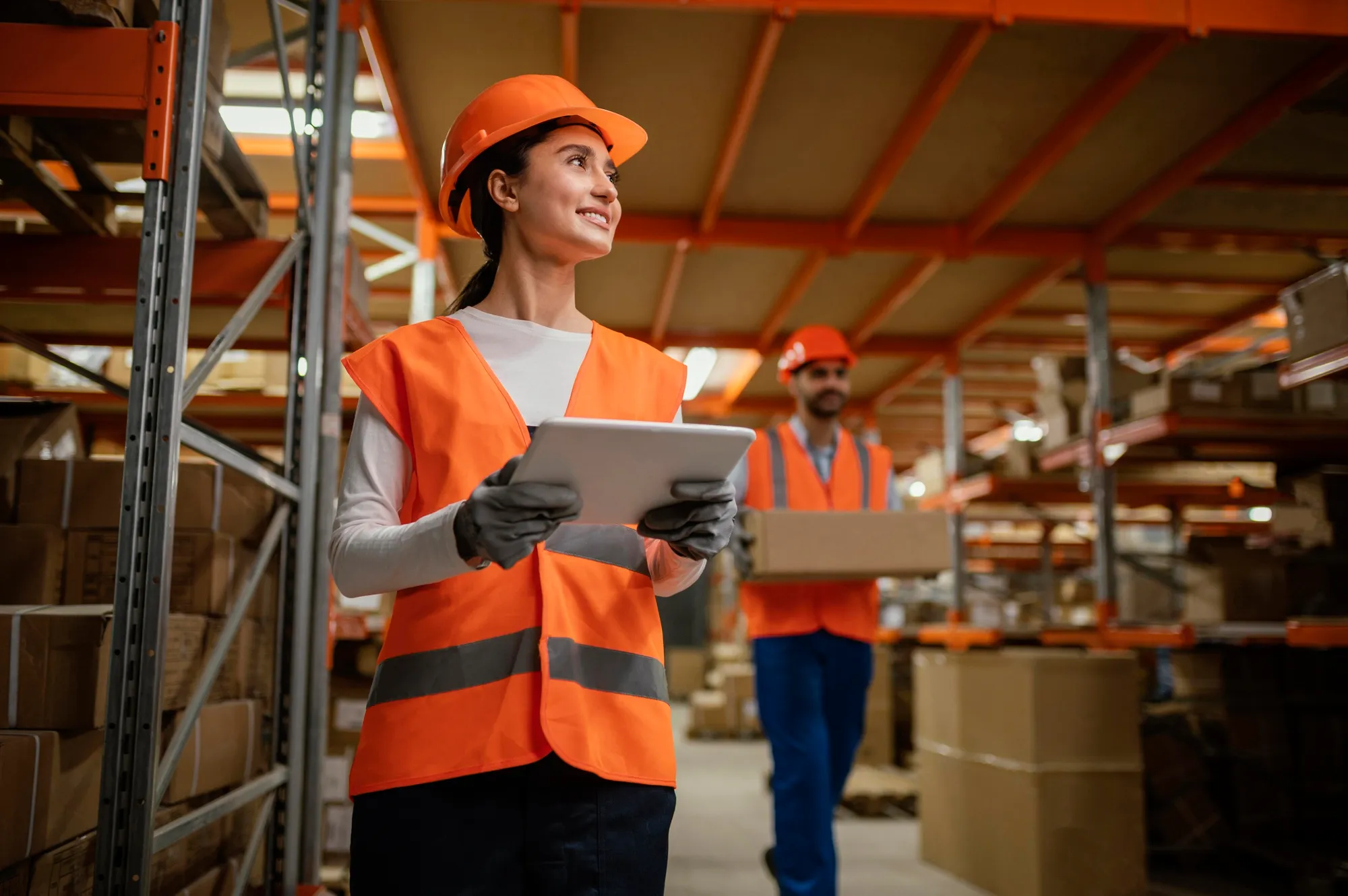 Woman in orange safety vest and helmet holding tablet in warehouse, man carrying box in background.